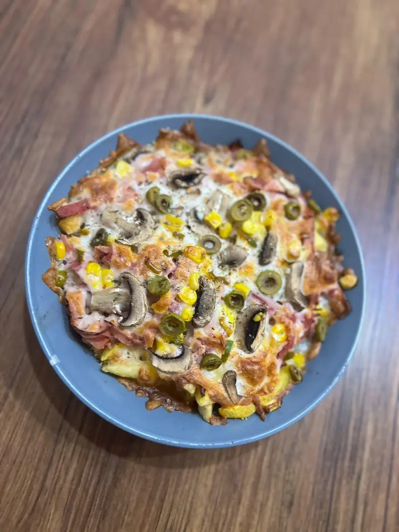 تصویر غذای خاص کافه پارس: پیتزا یا گراتن قارچ و ذرت با پنیر آب شده و زیتون، ظاهری طلایی و دلچسب در کاسه‌ای آبی روی میز چوبی.