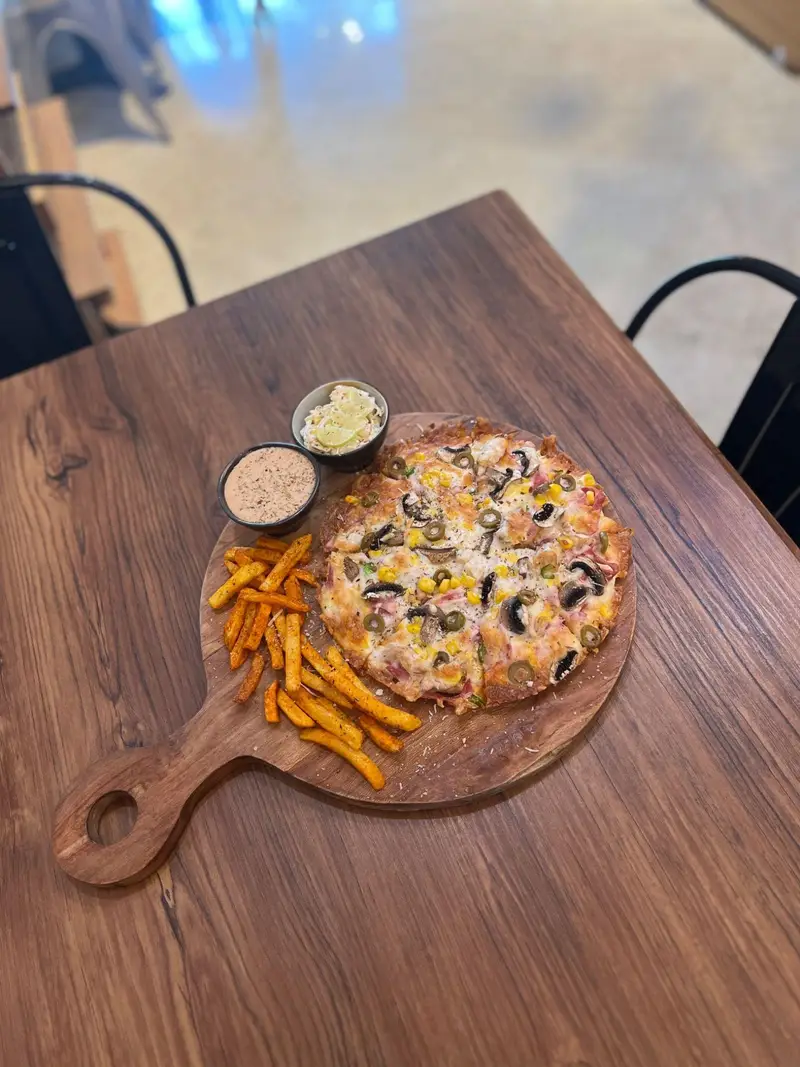 پیتزا قارچ و زیتون با سیب زمینی سرخ کرده و سس‌های مخصوص، یک غذای خوشمزه و کامل از منوی کافه پارس روی تخته چوبی.