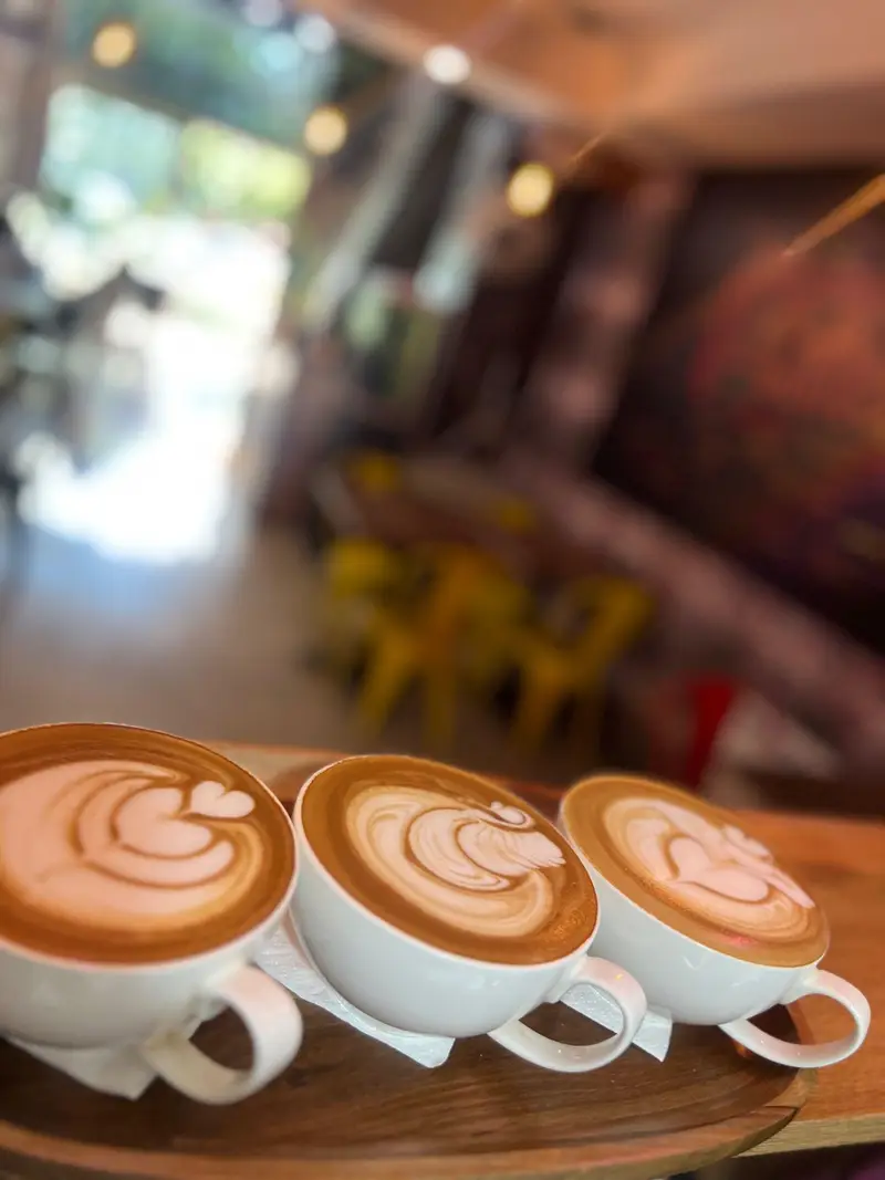 تصویری جذاب از سه فنجان قهوه لاته آرت خاص و بی‌نظیر کافه پارس روی سینی چوبی. لذت نوشیدنی گرم در فضایی صمیمی و دلپذیر.