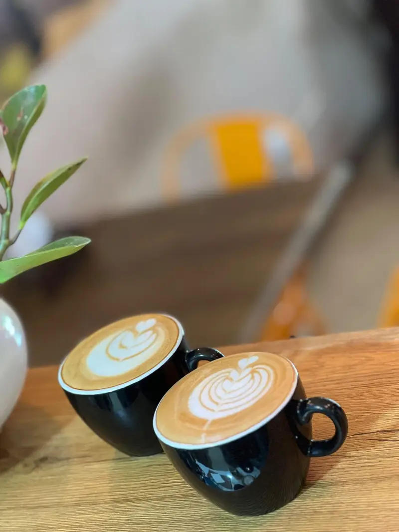 دو فنجان قهوه لاته آرت زیبا روی میز چوبی در کافه پارس، مکانی دنج و دلنشین برای نوشیدنی‌های گرم و لحظات آرامش‌بخش.