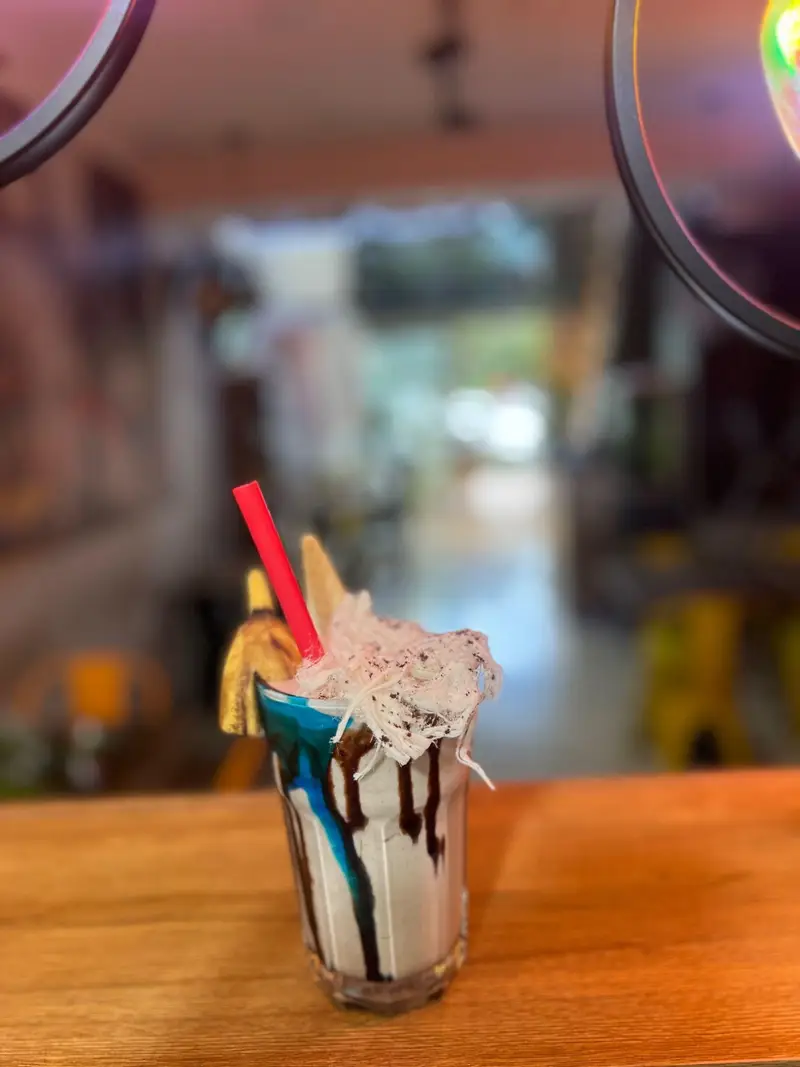 تصویری از یک میلک شیک منحصر به فرد کافه پارس، با سس شکلات، تزئین آبی و پشمک سفید، مناسب برای لحظات دلپذیر.