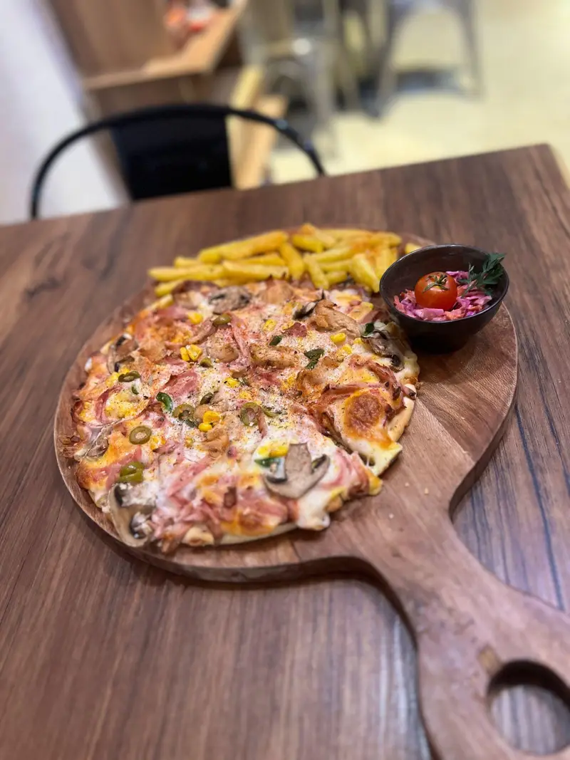 پیتزا مخصوص کافه پارس با قارچ، بیکن و ذرت، به همراه سیب‌زمینی سرخ کرده و سالاد تازه روی تخته چوبی. تجربه‌ای خوشمزه در کافه پارس.