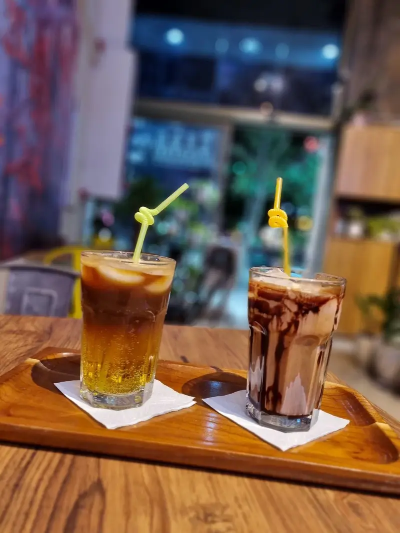 تصویری جذاب از دو نوشیدنی تابستانی کافه پارس: یک قهوه لایه ای و موکای شکلاتی با سس، روی سینی چوبی بر میز چوبی.