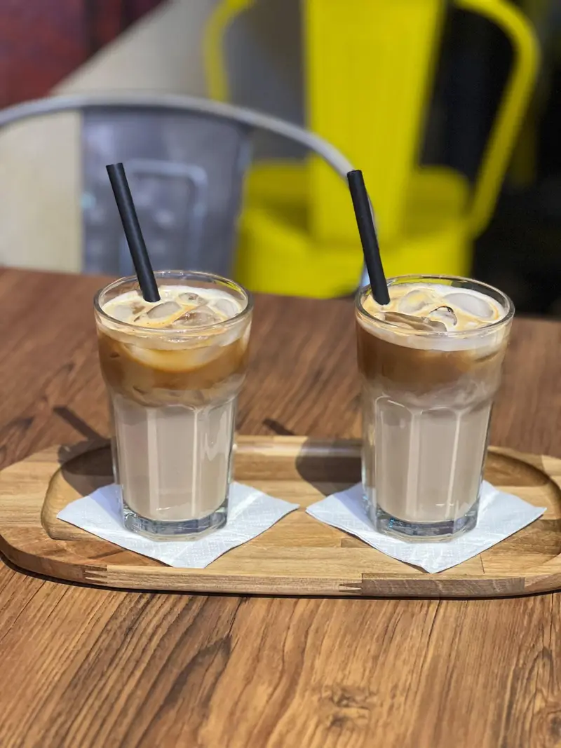 دو لیوان آیس لاته با نی مشکی در کافه پارس، به صورت لایه ای با یخ و شیر و قهوه سرو شده بر روی سینی چوبی روی میز چوبی.