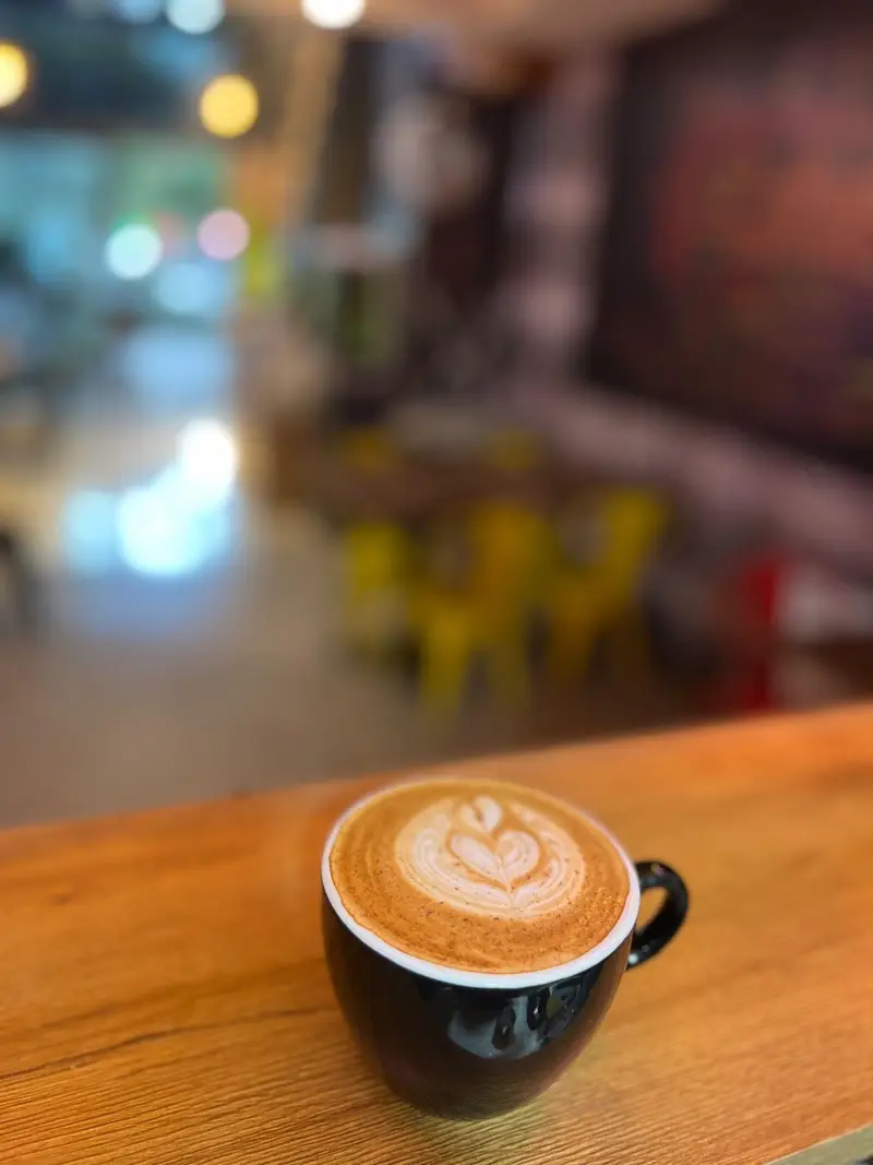 یک فنجان قهوه لاته با لاته آرت هنرمندانه به شکل قلب، روی میز چوبی روشن در فضای دلنشین کافه پارس، تجربه‌ای دلچسب از نوشیدنی‌های گرم.
