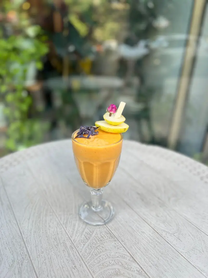 اسموتی پرتقالی تازه کافه پارس با تزئینات میوه و گل طبیعی، نوشیدنی خنک و سالم روی میز چوبی در فضای دلنشین کافه پارس.