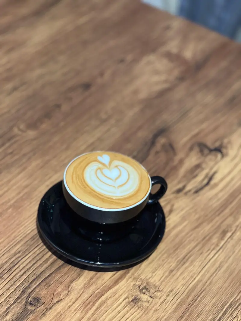 فنجان قهوه لاته با لاته آرت هنرمندانه طرح قلب روی میز چوبی در کافه پارس. لذت نوشیدن یک قهوه تازه و خوش عطر در فضایی دلنشین.