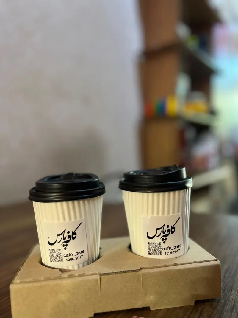 دو لیوان قهوه یکبار مصرف سفید با درپوش مشکی از کافه پارس در سینی حمل. شامل لوگو، QR کد و آدرس اینستاگرام. مناسب سفارش بیرون‌بر.