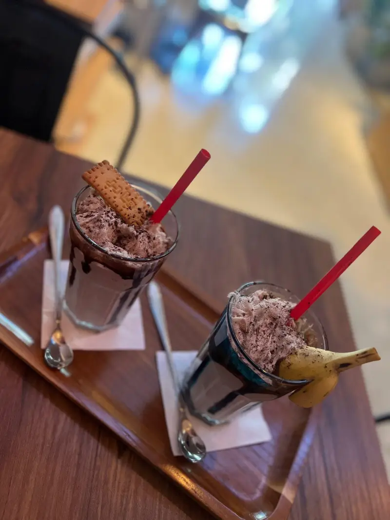 دو میلک شیک جذاب شکلاتی و موزی، با تزئینات خامه، پودر شکلات، بیسکوییت و موز تازه، سرو شده در سینی چوبی کافه پارس.