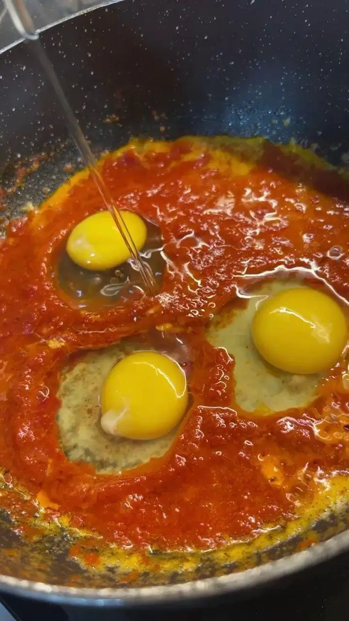 تخم مرغ‌های تازه در سس گوجه‌فرنگی قرمز و غلیظ در حال پخت در تابه، نمایی از آشپزی دلچسب و آماده‌سازی صبحانه یا املت خوشمزه در کافه پارس.
