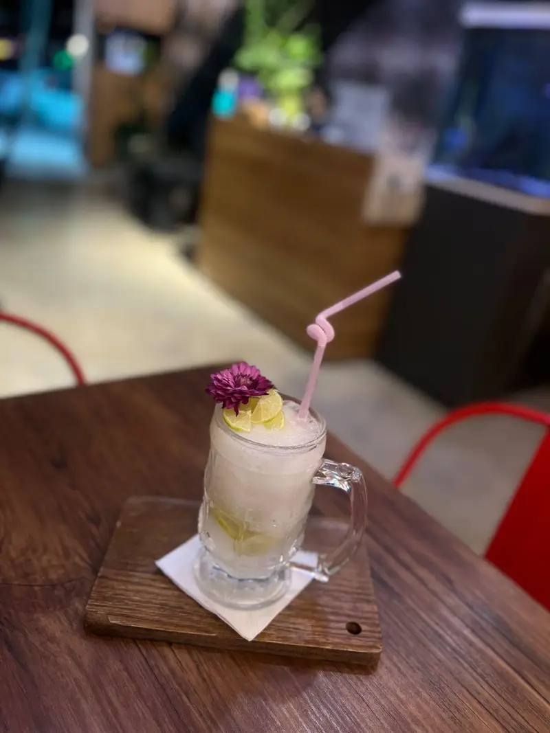 یک لیوان بزرگ نوشیدنی خنک و تازه لیمویی (احتمالاً لیموناد یا اسموتی) با تزیین لیمو و گل بنفش و نی صورتی روی میز چوبی کافه پارس.