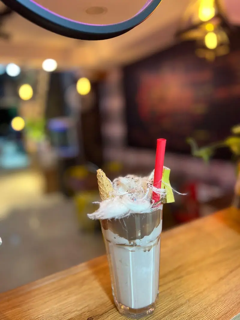 میلکشیک خوشمزه شکلاتی کافه پارس با پشمک رنگارنگ، قیف وافل و نی قرمز، سرو شده بر روی میز چوبی در محیط گرم و دلنشین کافه.