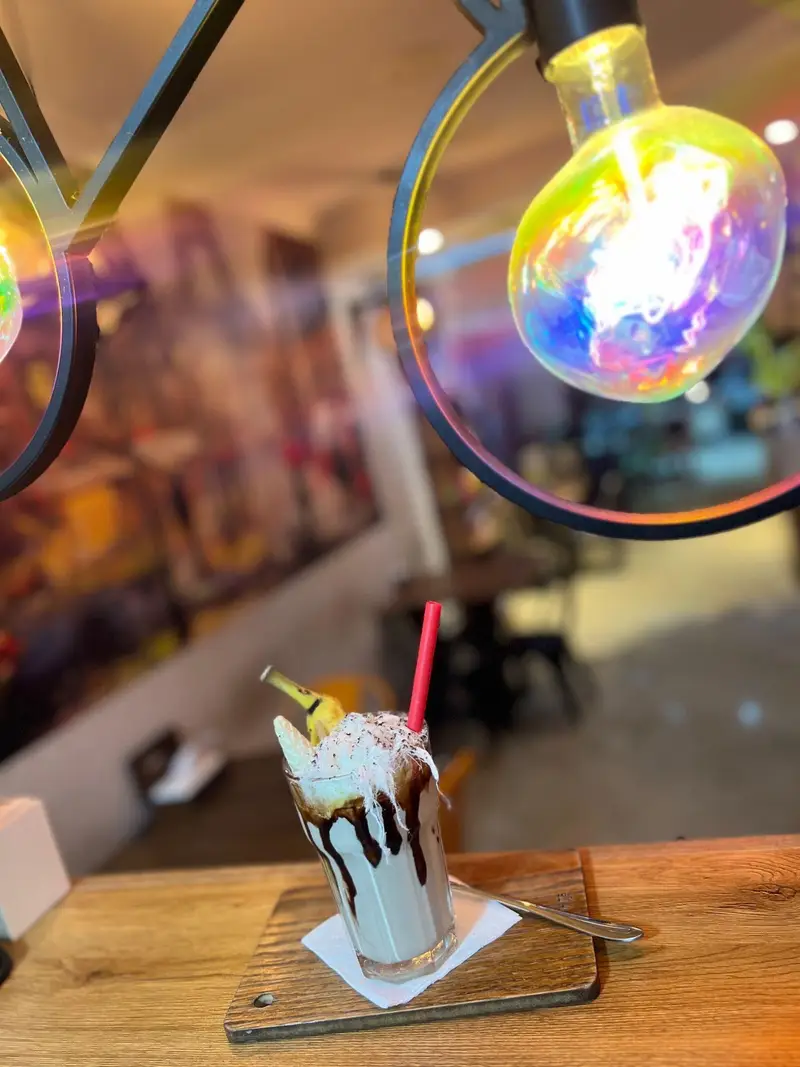 میلکشیک خوشمزه و خنک کافه پارس با تزئین خامه، سس شکلات و موز، سرو شده روی میز چوبی در فضای گرم و دلنشین کافه