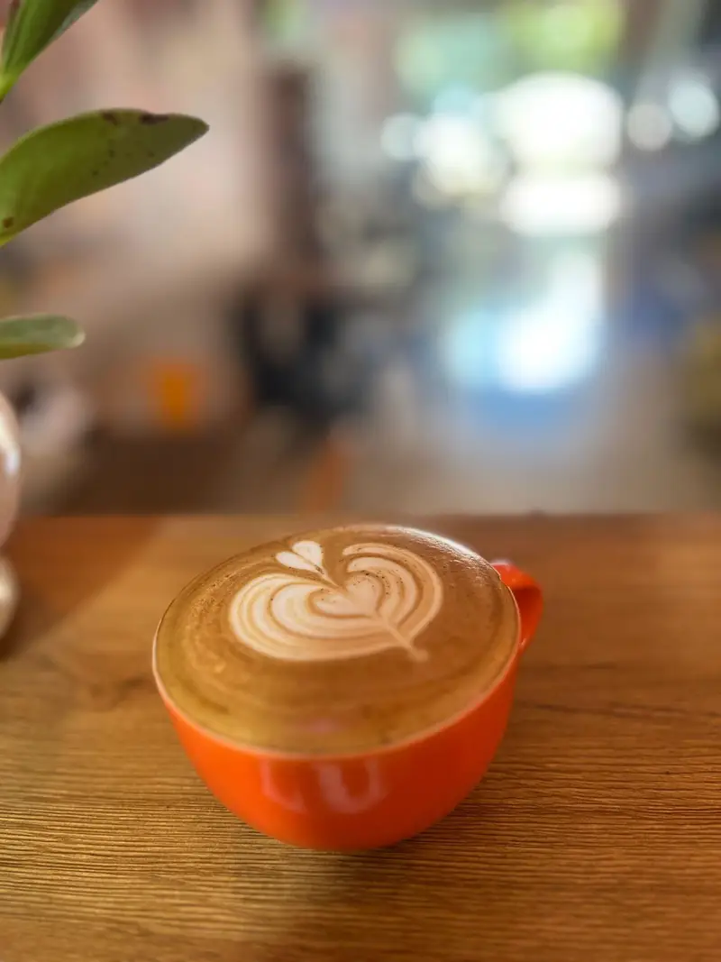 لاته آرت با طرح قلب در فنجان نارنجی بر روی میز چوبی کافه پارس. تجربه نوشیدنی گرم در محیطی دلنشین و آرام.