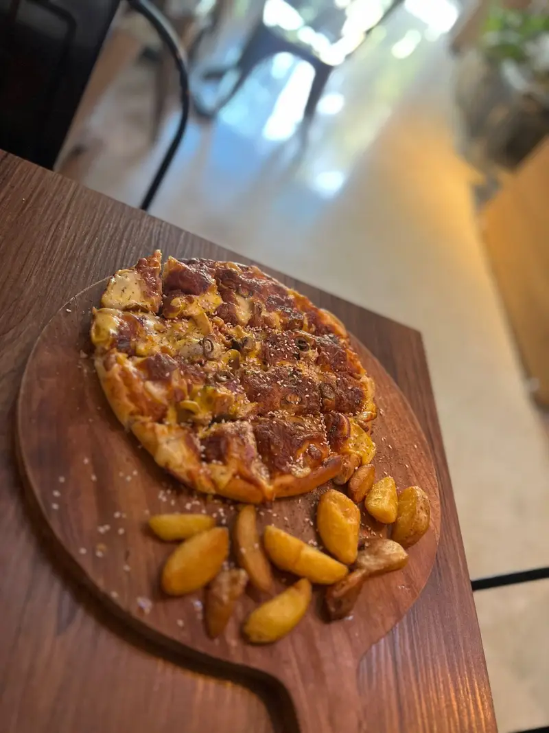 یک پیتزای پنیری خوشرنگ و طلایی با برش‌های آماده سرو، همراه سیب‌زمینی تنوری و چاشنی نمک، بر روی تخته چوبی در کافه پارس.