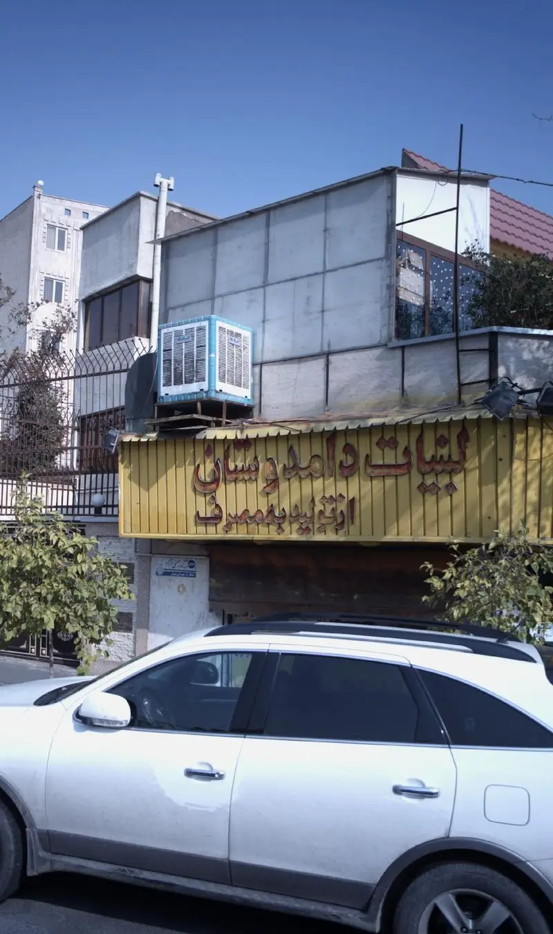 تصویری از نمای خارجی کسب و کار لبنیات دوستان، شامل تابلوی زرد رنگ، کولر آبی روی پشت بام و یک خودروی شاسی بلند سفید در خیابان.