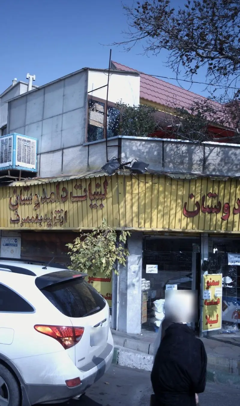 نمای بیرونی مغازه لبنیات دوستان با تابلوی زرد رنگ، نوشته فارسی "از تولید به مصرف"، خودروی سفید و رهگذر در خیابان.