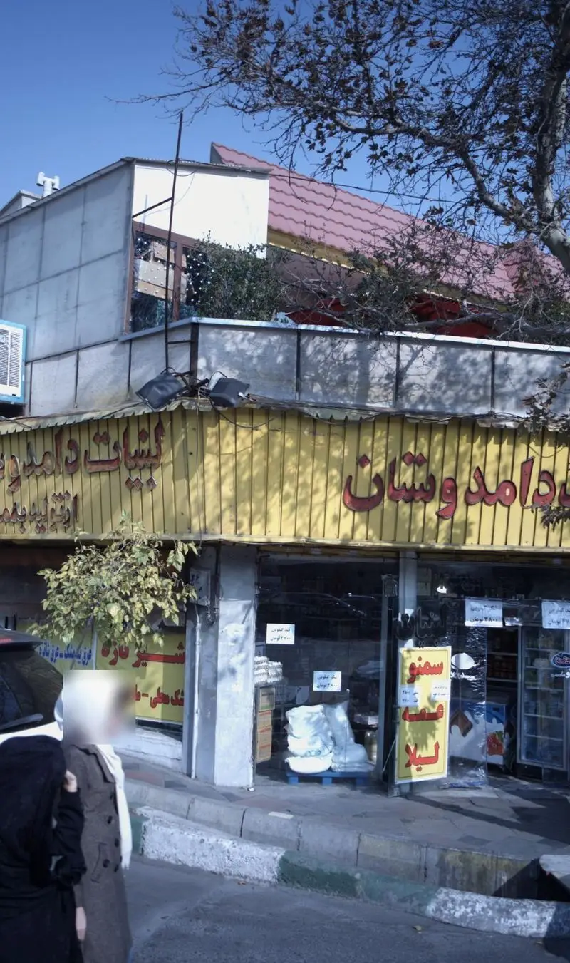 تصویر نمای خارجی مغازه لبنیات دوستان با تابلوی زرد روشن، نمایش محصولات لبنی در ویترین و زندگی روزمره در خیابان شلوغ.