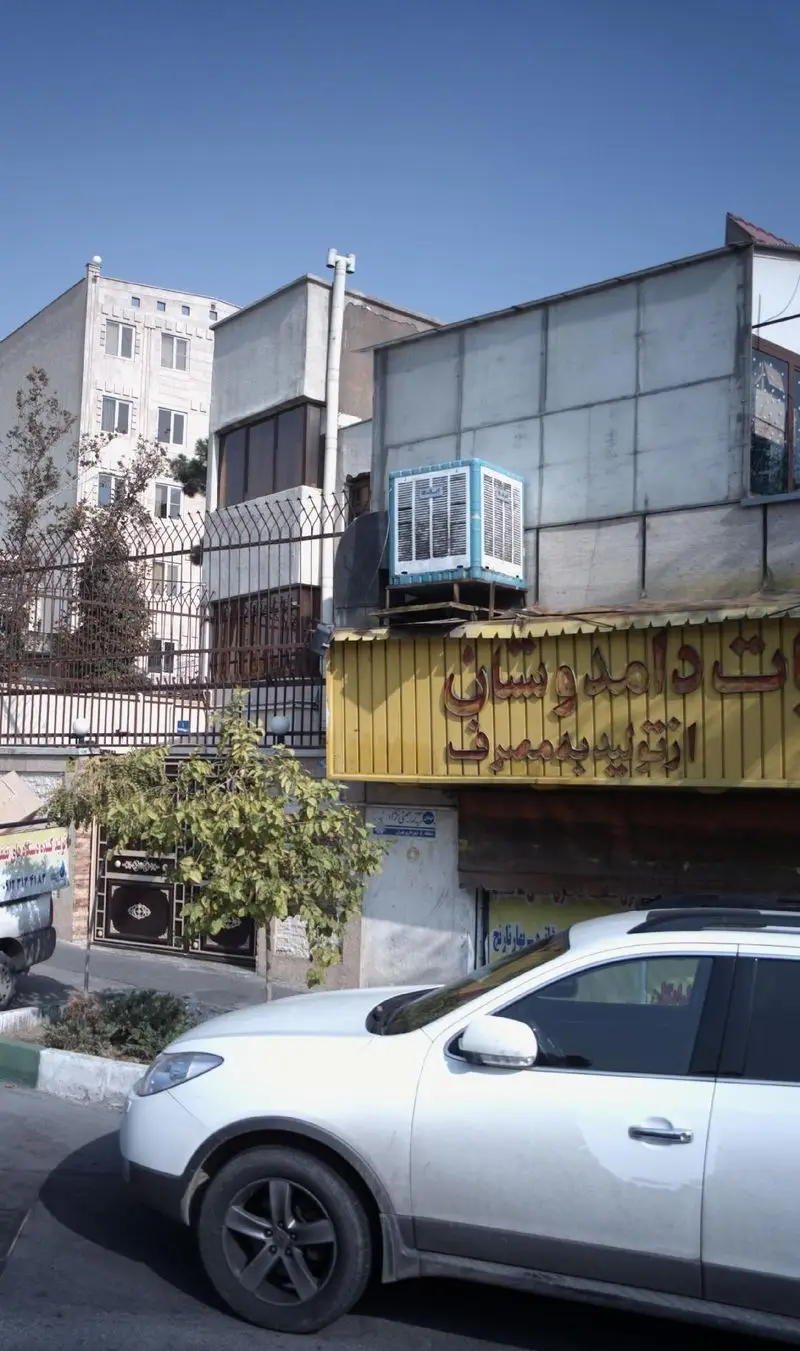 تصویر نمای بیرونی فروشگاه لبنیات دوستان با تابلوی زرد رنگ و برجسته، کولر آبی روی سقف و یک خودروی سفید پارک شده در خیابان.