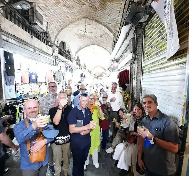 گروهی از گردشگران شاد در حال لذت بردن از نوشیدنی‌ها در فضای سنتی بازار، مرتبط با قهوه خانه حاج علی درویش.