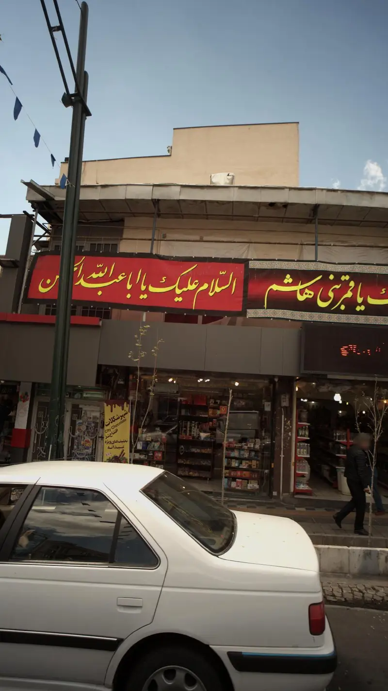 نمای بیرونی فروشگاه شهر شکلات قلهک در روز، همراه با بنرهای مذهبی بزرگ، ویترین محصولات و خودروی سفید رنگ در خیابان.