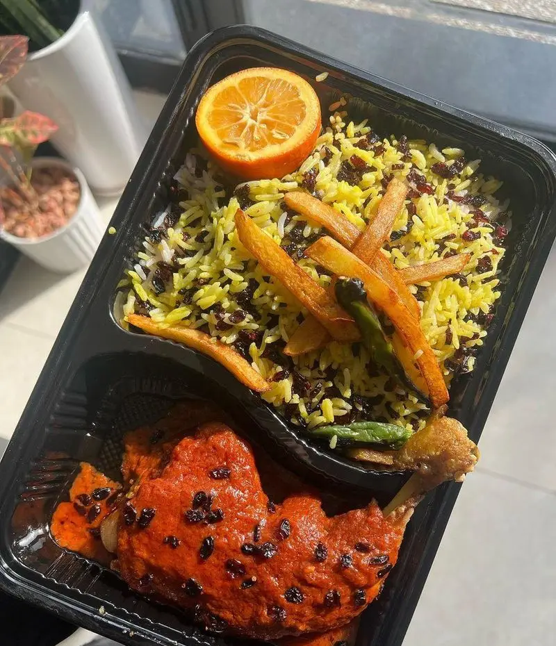 یک پرس غذای ایرانی زرشک پلو با مرغ از رستوران ضیافت، شامل برنج زعفرانی، مرغ ران با سس قرمز، زرشک، سیب زمینی و پرتقال در ظرف بیرون‌بر.