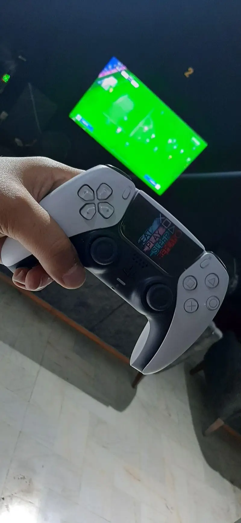 یک دست در حال نگه داشتن کنترلر سفارشی DualSense PS5 با متن "EAT PLAY SLEEP REPEAT" در گیم‌نت VIP PS5 پارس، با صفحه نمایش بازی فوتبال در پس زمینه.