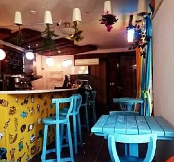 فضای داخلی کافه آرام با دکوراسیون رنگارنگ، شامل میز و صندلی‌های آبی، بار زرد و سقف خلاقانه با گلدان‌های وارونه و نورپردازی جذاب.