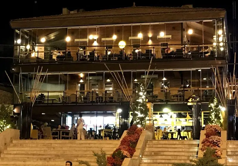 نمایی از کافه رستوران پلاس ویونا در شب، با دو طبقه شیشه‌ای، نورپردازی گرم، دکوراسیون چوبی و مشتریان در فضای دلنشین.