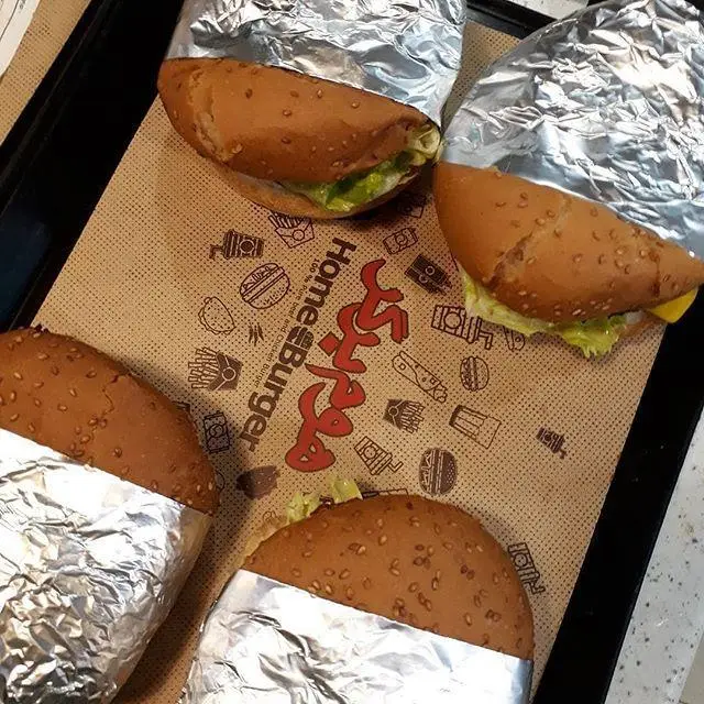 تصویری از چهار ساندویچ برگر خوشمزه هوم برگر با نان کنجدی، کاهو و بسته بندی فویل آلومینیومی روی کاغذ برند.