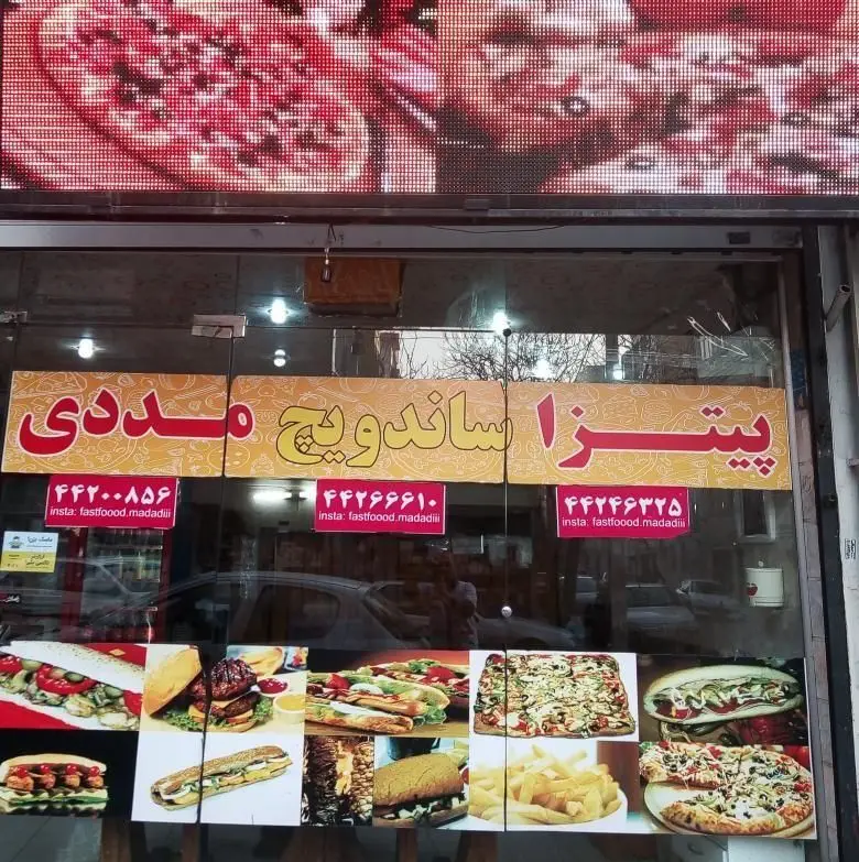 نمای بیرونی پیتزا و ساندویچ مددی، با تابلو و منوی متنوع شامل پیتزا، ساندویچ، برگر و سیب زمینی، اطلاعات تماس و اینستاگرام.