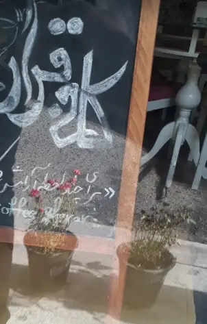 تخته سیاه تبلیغاتی کافه قرار با نام، شعار و نوشته Coffee Gharar، ورودی جذاب کافه با گلدان‌های زیبا.