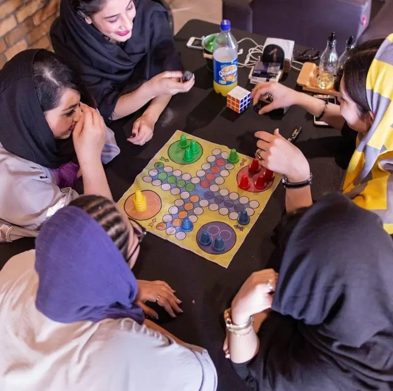 زنان شاد در کافه خیابون، با روسری‌های رنگی، در حال بازی رومیزی هیجان‌انگیز و دورهمی دوستانه. تجربه‌ی لحظات لذت‌بخش و سرگرمی.