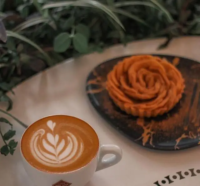 تصویری از یک فنجان لاته با لاته آرت جذاب و دسر رز شکل خوشمزه در کافه فلورین. لحظاتی دلنشین با قهوه‌های تازه و شیرینی‌های خاص.