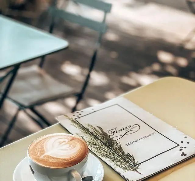 یک فنجان لاته با لاته آرت قلب، در کنار منوی کافه و رستوران ایتالیایی فلورین با تزئین رزماری، فضایی دلنشین و آرامش‌بخش.