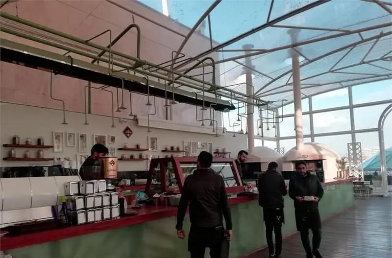 کافه رستوران فلورین: نمای داخلی با سقف شفاف، دو کوره پیتزا، بار سرویس نوشیدنی و غذا، و پرسنل فعال در فضایی دلباز.