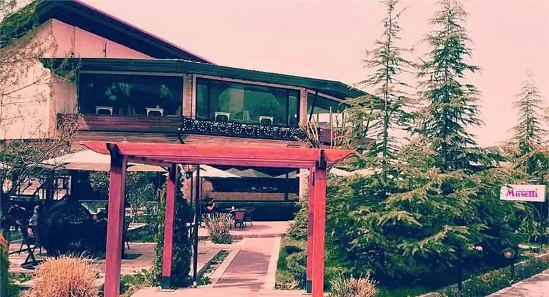 کافی شاپ بورا بورا: نمایی جذاب از ساختمان اصلی با تراس شیشه‌ای، محوطه بیرونی زیبا شامل میز و صندلی‌ها، آلاچیق‌های چوبی قرمز و درختان سبز.