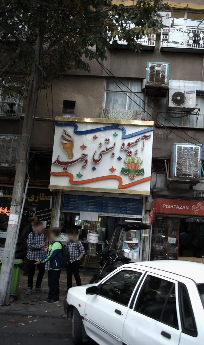 نمایی از تابلو بزرگ و رنگی آبمیوه بستنی وحید روی سردر مغازه، با طرح بستنی و هویج، در یک روز آفتابی کنار خیابان شلوغ شهر.