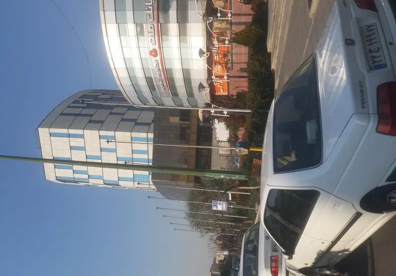 خیابان شهری با خودروهای پارک شده (پژو سفید)، ساختمان‌های مدرن بانکی و تجاری با نمای شیشه‌ای و آسمان آبی. محیطی شلوغ در دل شهر.