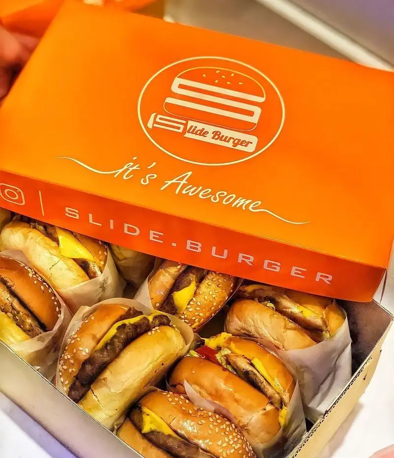 تصویر جعبه نارنجی برند Slide Burger پر از مینی همبرگرهای لذیذ و داغ با نان کنجدی، آماده برای یک وعده غذایی عالی و جذاب.