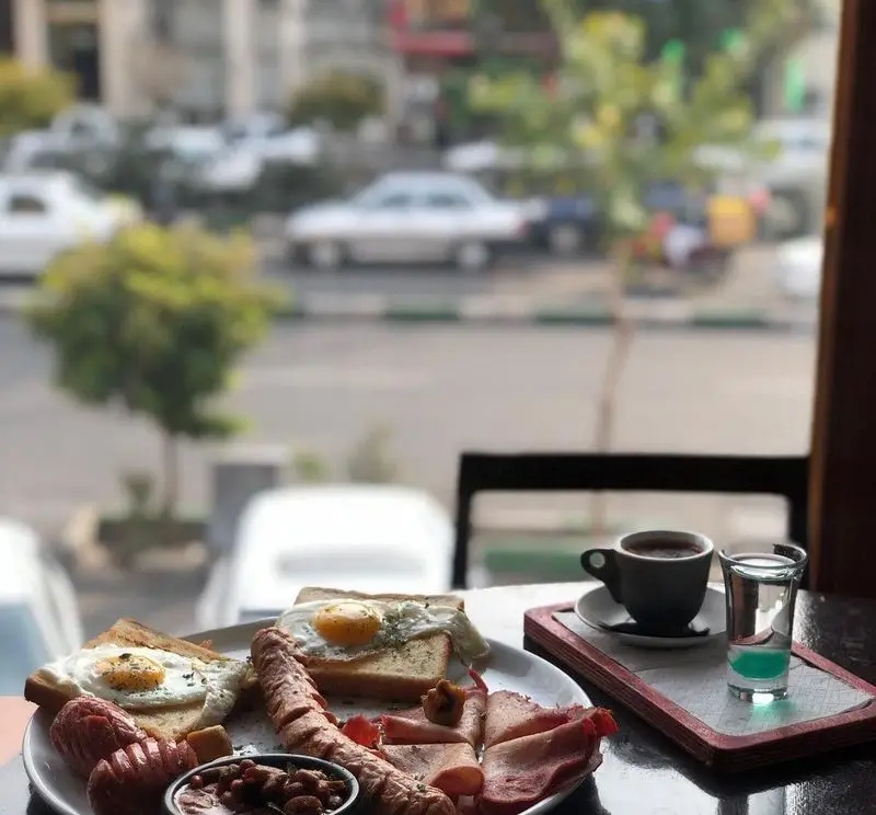 صبحانه انگلیسی کامل و لذیذ در رستوران ایتالیایی ژولی شامل تخم مرغ، سوسیس، ژامبون، لوبیا و قهوه با نمای شهر.