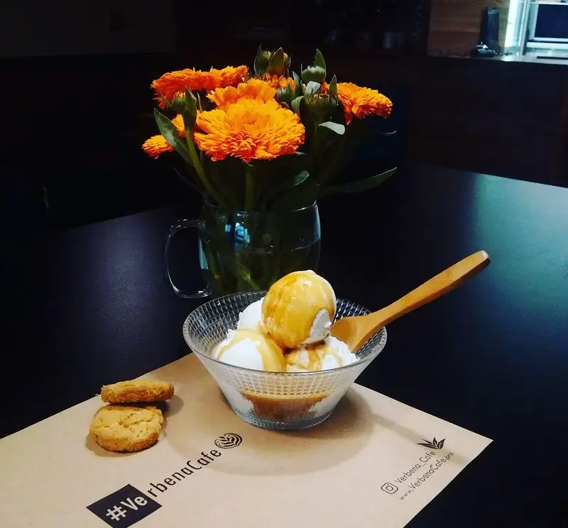 دسر آفوگاتو لذیذ کافه وربنا: بستنی وانیلی، سس کارامل و کلوچه تازه، در محیطی دلنشین با گل‌های نارنجی.