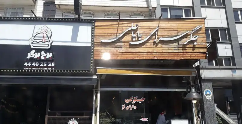 تصویر نمای بیرونی جگرسرای بابا علی، رستوران جگرکی با تابلوی چوبی شیک و نام فارسی برجسته. در کنار آن برج برگر نیز دیده می‌شود.