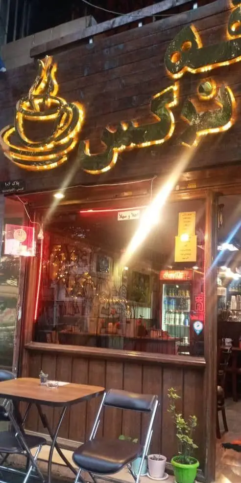 نمای بیرونی کافه اثر در شب با تابلوی درخشان و لوگوی فنجان قهوه، میز و صندلی‌های فضای باز، منعکس کننده محیط دنج داخلی.