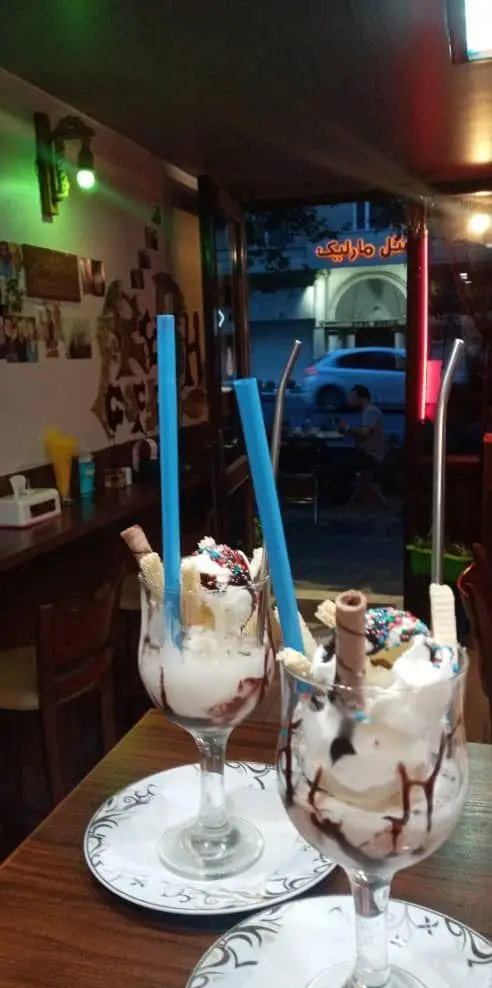 تصویری از دو لیوان بستنی دسر خوشمزه و تزئین شده با خامه، سس شکلات و ویفر در کافه اثر. فضایی آرامش‌بخش برای لذت بردن از طعم‌های دلنشین.