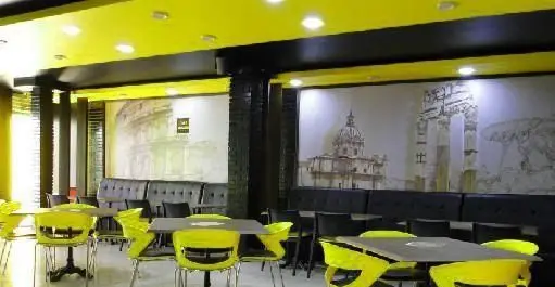 فضای داخلی مدرن فست فود آماندا با سقف زرد روشن، صندلی‌های مشکی و زرد، میزهای غذاخوری و دیوارنگاره‌های جذاب بناهای رومی.