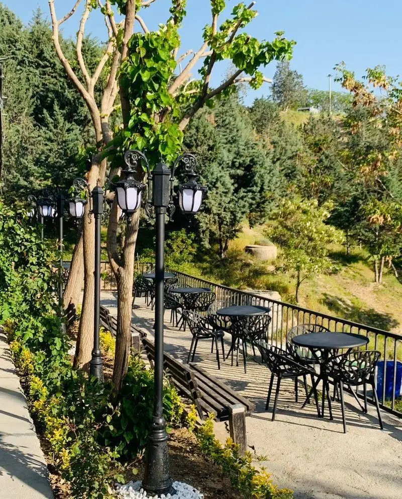 نمایی از فضای باز کافه اسکواش برج میلاد، شامل میز و صندلی‌های شیک مشکی، چراغ‌های قدیمی و درختان سرسبز در فضایی دنج و دلنشین.