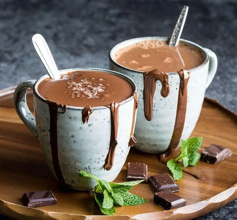 شکلات داغ کرمی و لذیذ کافه باران با سس شکلات روی ماگ‌های زیبا، همراه با تکه‌های شکلات و برگ‌های نعنا، تجربه‌ای بی‌نظیر.