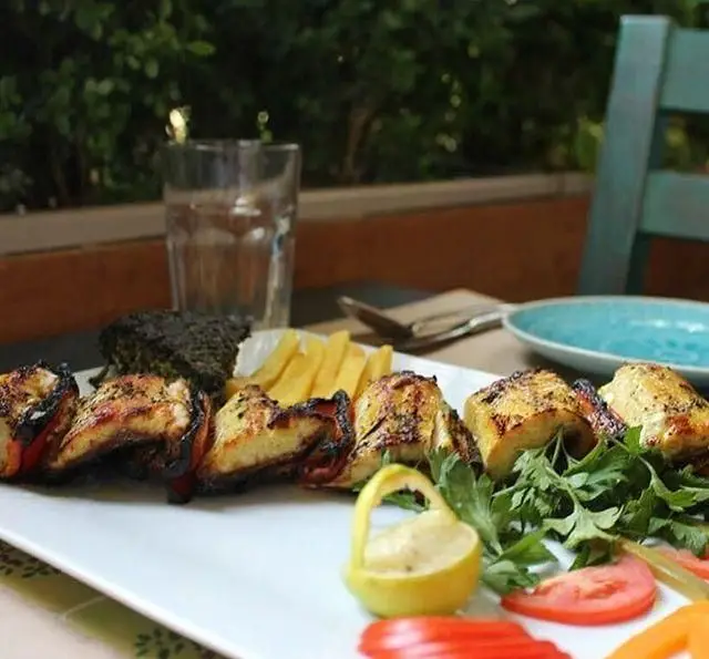 سفره ای دلنشین از جوجه کباب با سیب زمینی سرخ کرده، سبزی تازه و چاشنی لیمو در ایوان رستوران؛ بهترین طعم ها در فضای سبز.