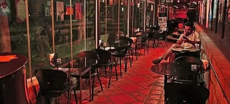 نمایی از محیط دنج و هنری کافه خانه هنرمندان تهران در شب با میز و صندلی‌های مدرن و مشتری در حال استراحت و استفاده از موبایل.